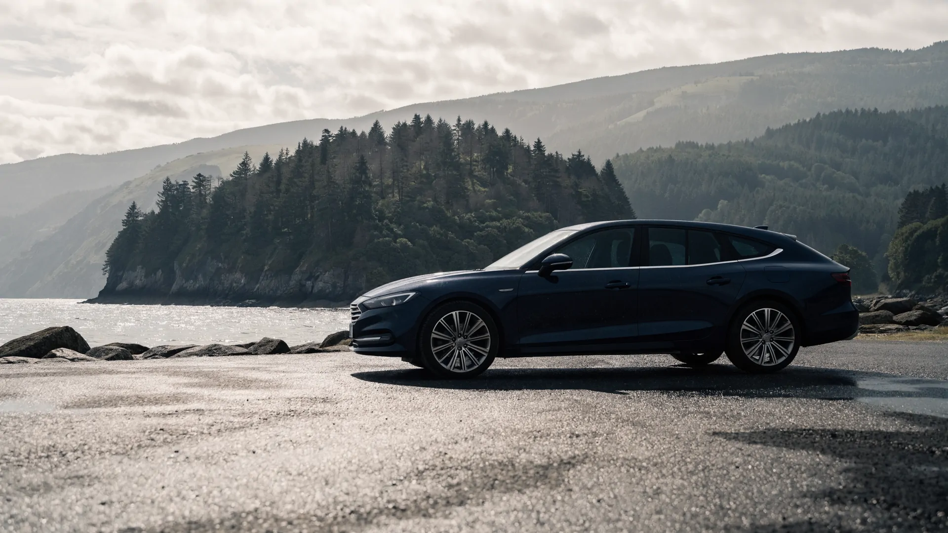 A navy estate car at a quiet coastal turnout, with water and evergreen hills beyond